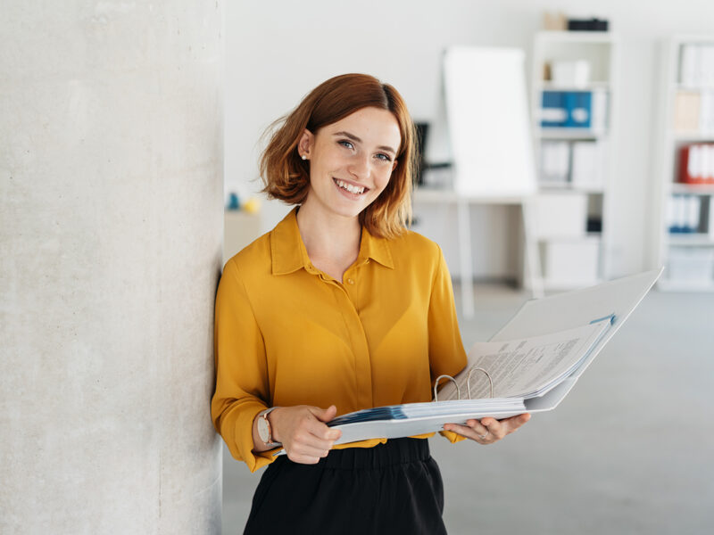Attractive young office worker holding a large open binder as she looks at the camera with a sweet friendly smile